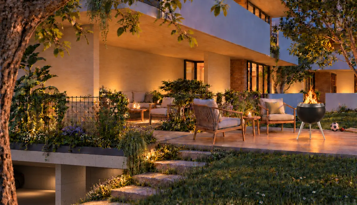 Das Haus am Abend von der Wiese aus: auf der Terrasse brennt eine Feuerschale zwischen zwei Sesseln, dahinter der warm erleuchtete Wohnbereich hinter bepflanzten Brüstungen; links führen Stufen hinab, darunter liegt der Stellplatz, im Vordergrund ein alter Baum.