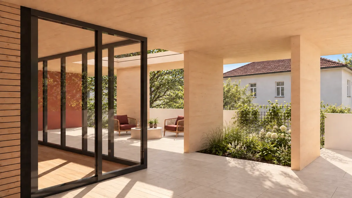 Eine überdeckte Terrasse unter der Decke aus Brettsperrholz: links die dunkel gerahmte Schiebetürwand zum Wohnraum, hinter ihr ein Hof mit rot verputzter Wand, zwei roten Sesseln und einem runden Steintisch unter einem Baum; rechts ein Beet mit Gräsern und weißen Hortensien hinter einem Gitter, dahinter das Nachbarhaus mit Ziegeldach.