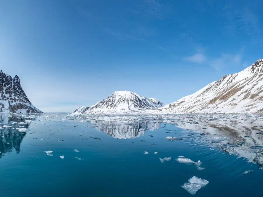 Ein Fjord im Polarsommer: stilles, tiefblaues Wasser mit treibenden Eisstücken, ringsum verschneite Berge, darüber ein klarer Himmel.