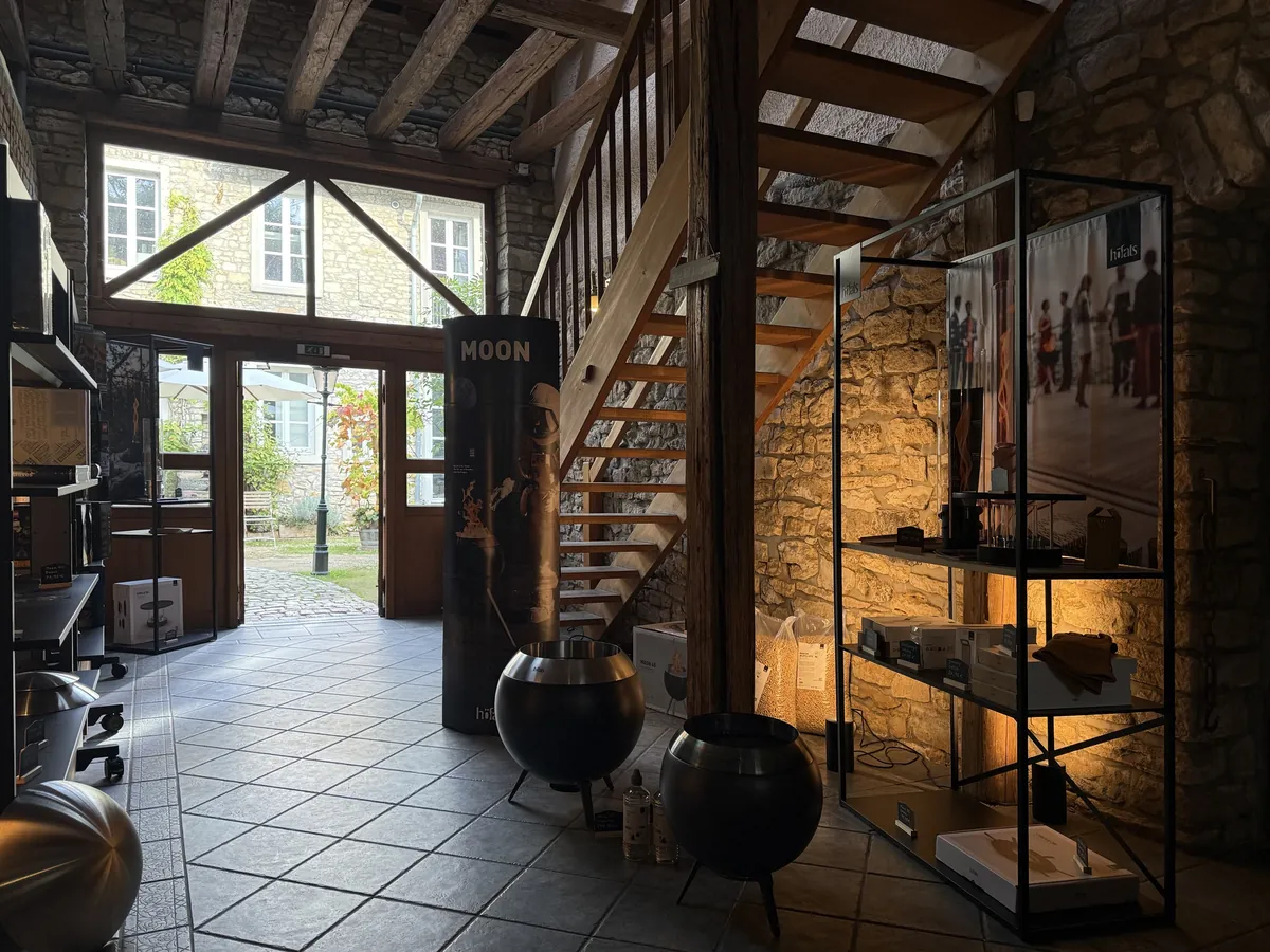 Blick in den Laden: unter alten Holzbalken und einer offenen Holztreppe stehen runde Feuerschalen aus Stahl und schwarze Regale vor einer warm beleuchteten Bruchsteinwand; durch die offene Tür und das große Fenster sieht man in den Hof.