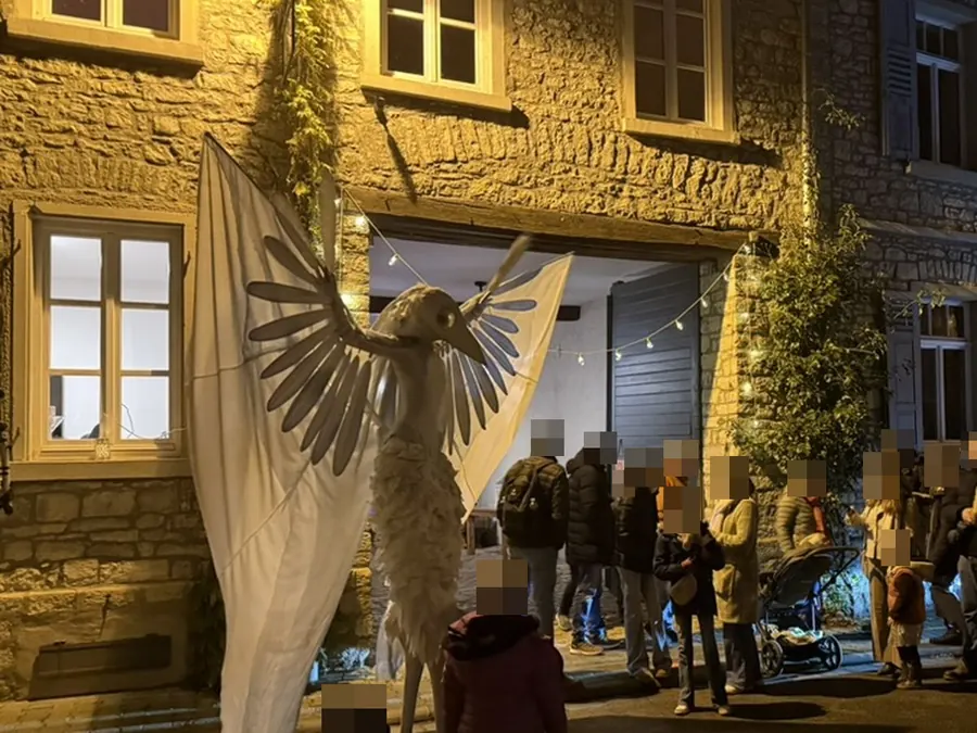 Abends in der Gasse: eine beleuchtete geflügelte Figur vor der Bruchsteinfassade, Zuschauer stehen im Halbkreis