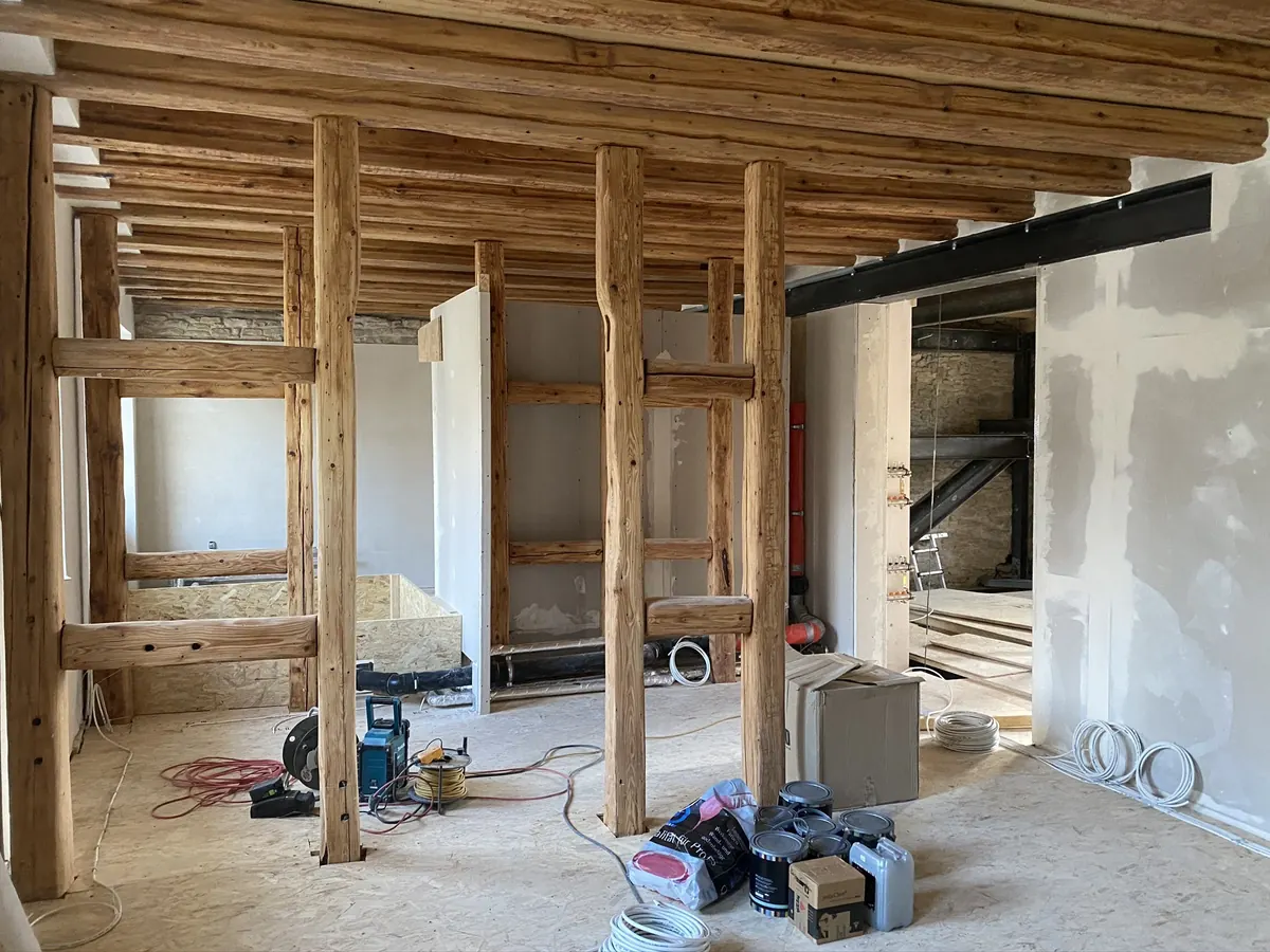 Ein Bestandsbau im Umbau: freigelegte Rundhölzer tragen als Stützen und Riegel die alte Balkendecke, dahinter stehen neue Wände; auf dem Boden liegen Kabelrollen, Farbeimer und Werkzeug.