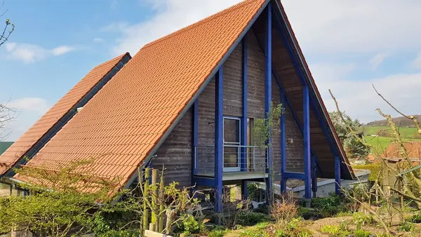 Ein Dach bis fast zum Boden — Blaue Stützen tragen die offene Ebene vor der Holzfassade.